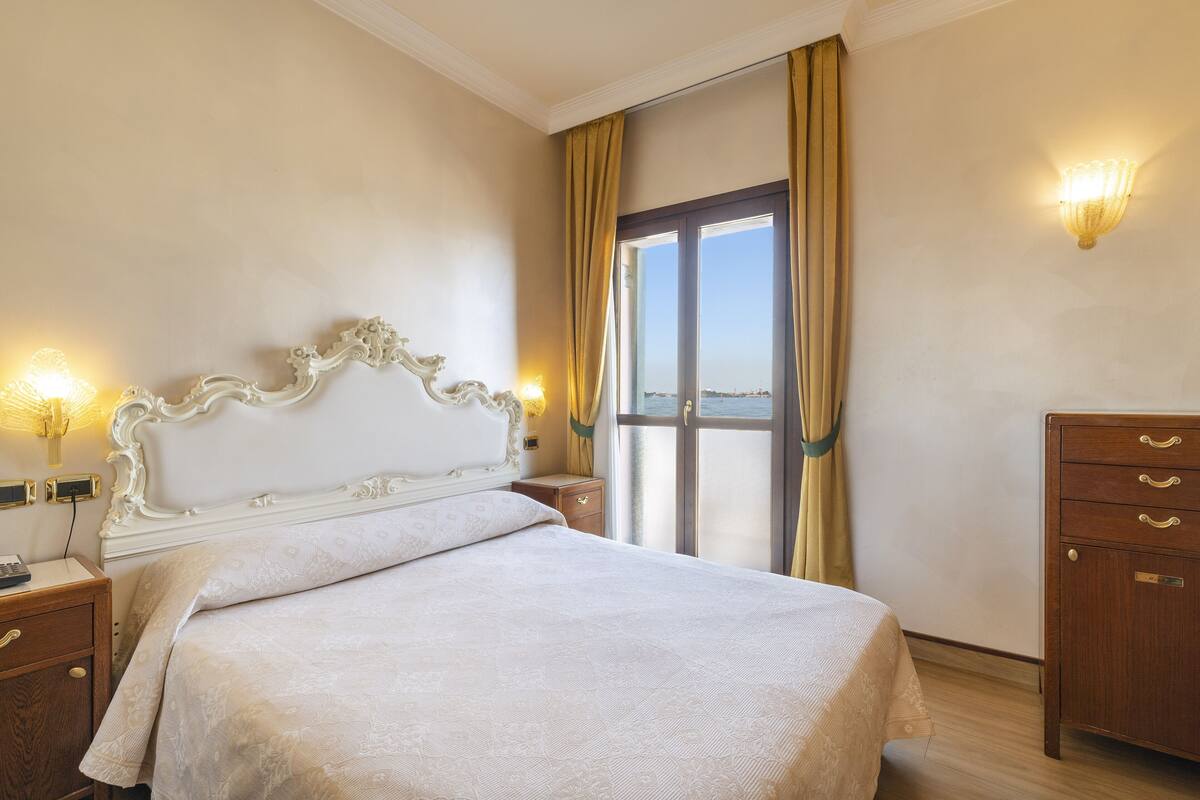 A hotel room with a large bed, a wooden dresser, a nightstand with a lamp, and a view of the sea through a large window.