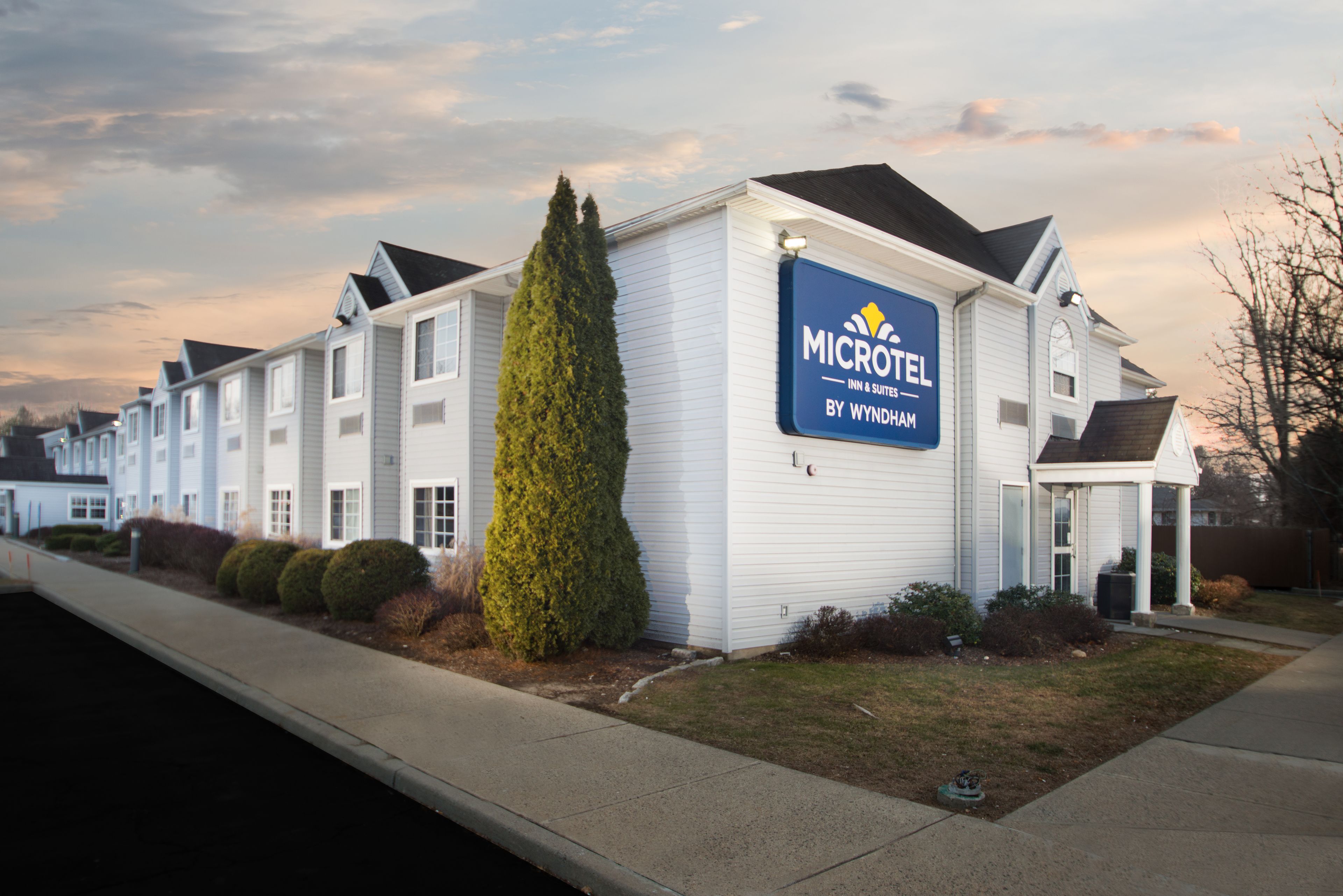 A Microtel Inn & Suites by Wyndham building with a white exterior, a large evergreen tree, and a clear sky.