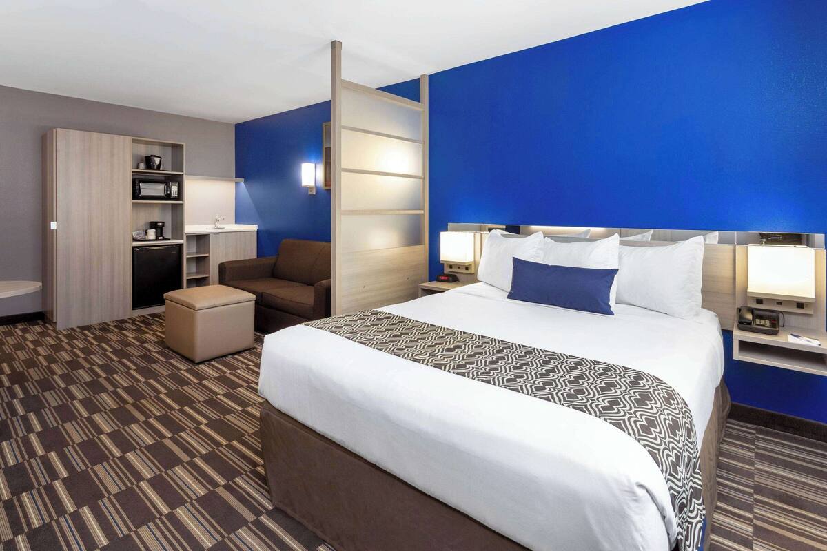 A hotel room with a large bed, a desk with a computer, a chair, a small kitchenette, and a bathroom.