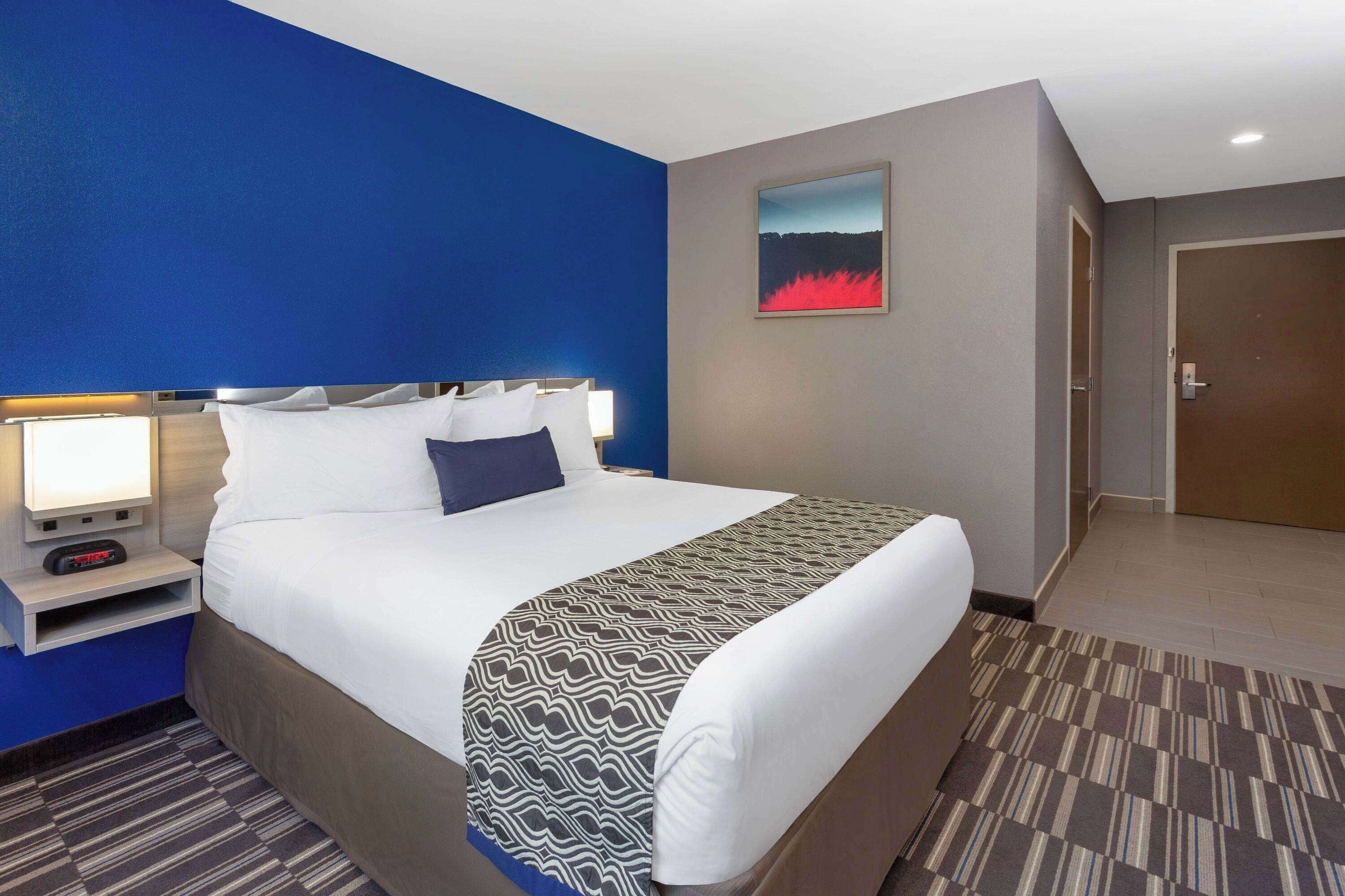A hotel room with a large bed, a nightstand with a lamp, a painting on the wall, and a door.