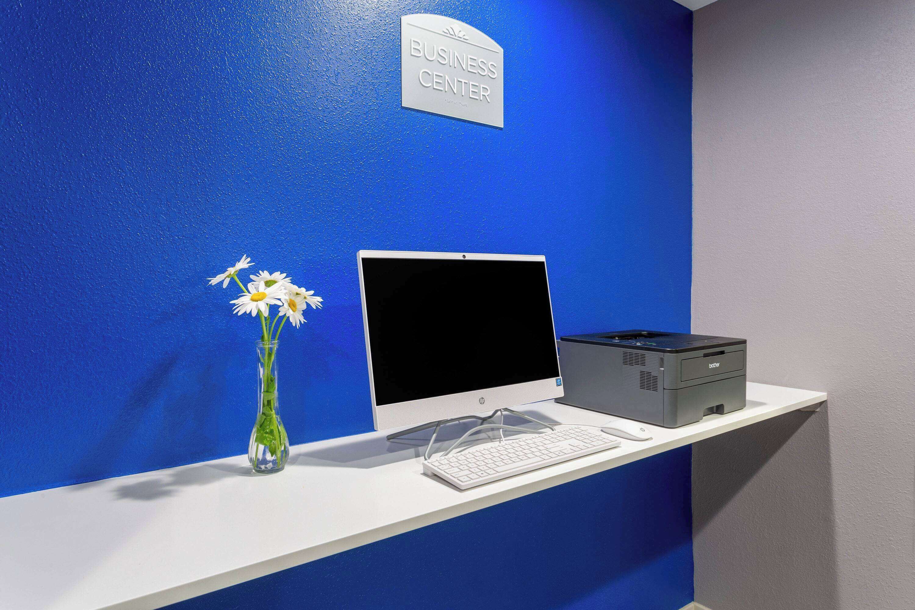 A business center with a desktop computer, a printer, and a vase with flowers on a white desk against a blue wall.