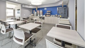 Un comedor moderno con mesas y sillas blancas, una barra con máquina de café y una pared azul decorativa.