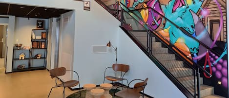 Un interior moderno con un mural de grafiti vibrante, una escalera con barandilla de malla y una zona de estar con sillas y una pequeña mesa.