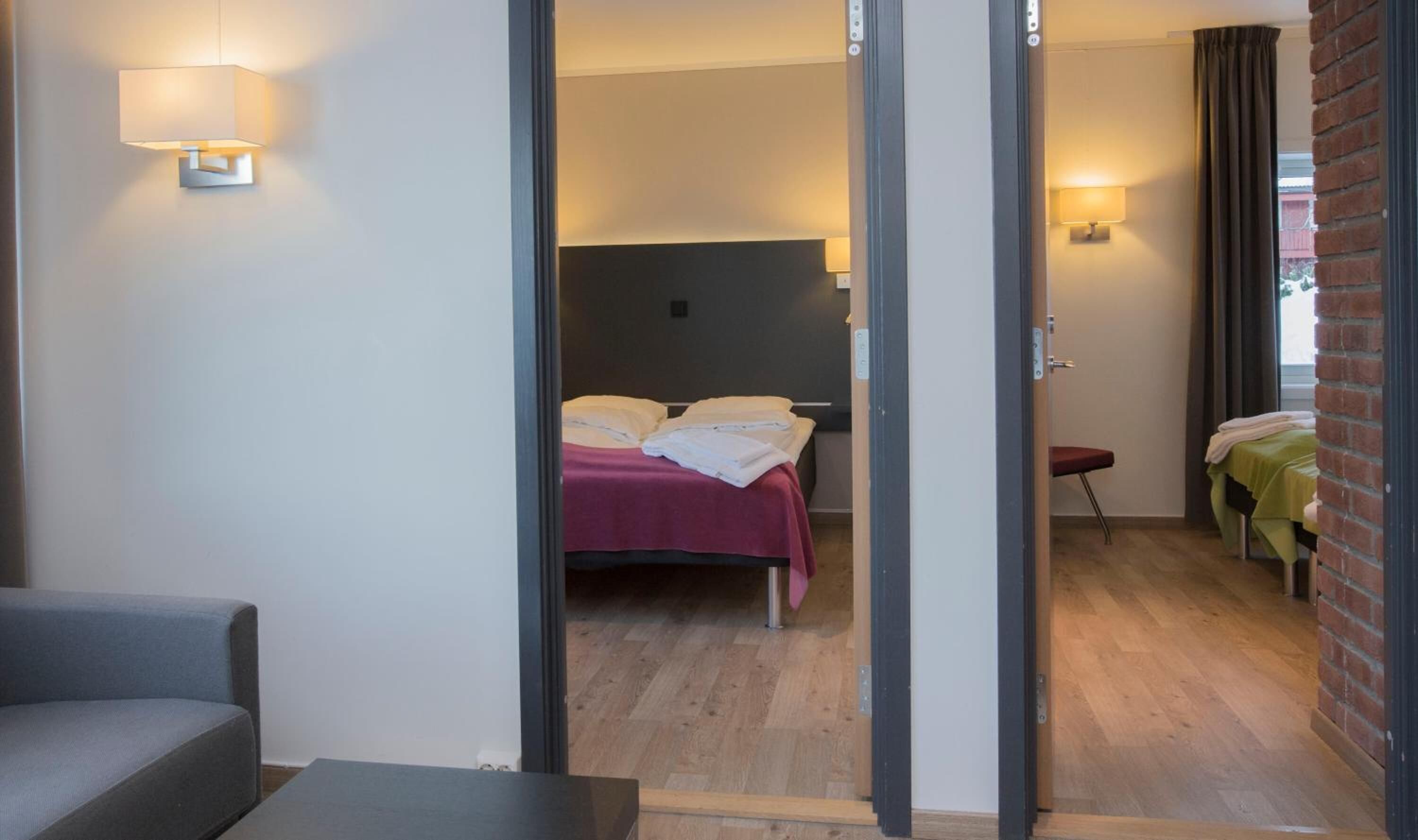 Habitación de hotel con una cama, una mesita y una silla. Incluye un baño con ducha y retrete. El piso es de madera y la pared, de ladrillo.