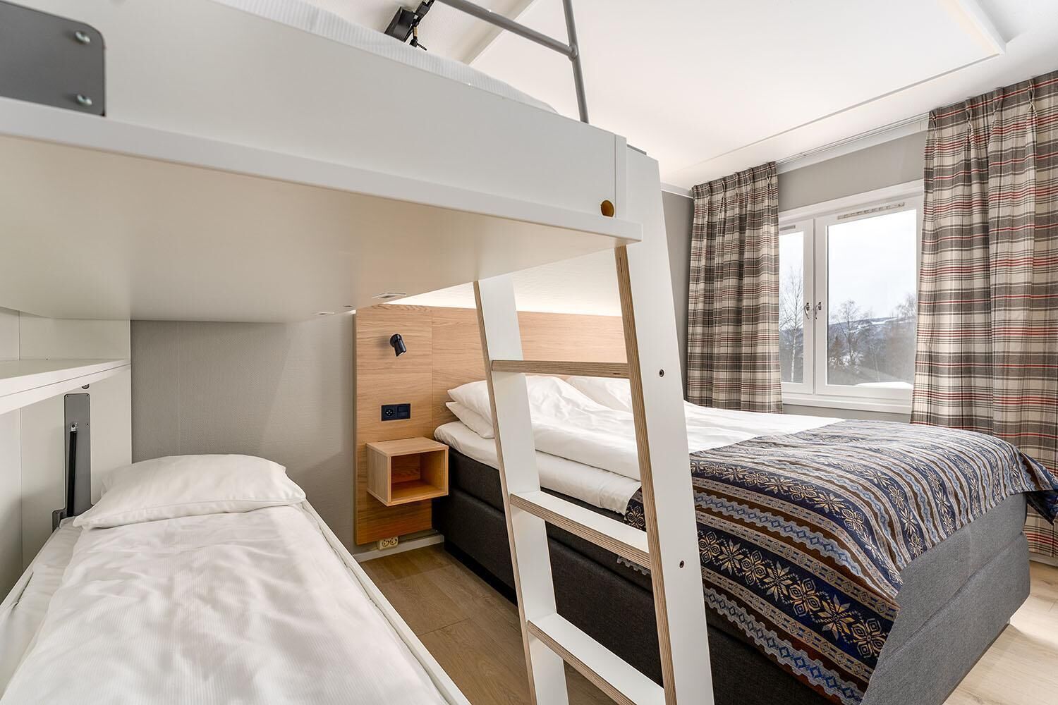 A compact hotel room with a bunk bed, a small bedside table, a window with curtains, and a patterned bedspread.