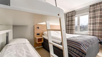 A compact hotel room with a bunk bed, a small bedside table, a window with curtains, and a patterned bedspread.