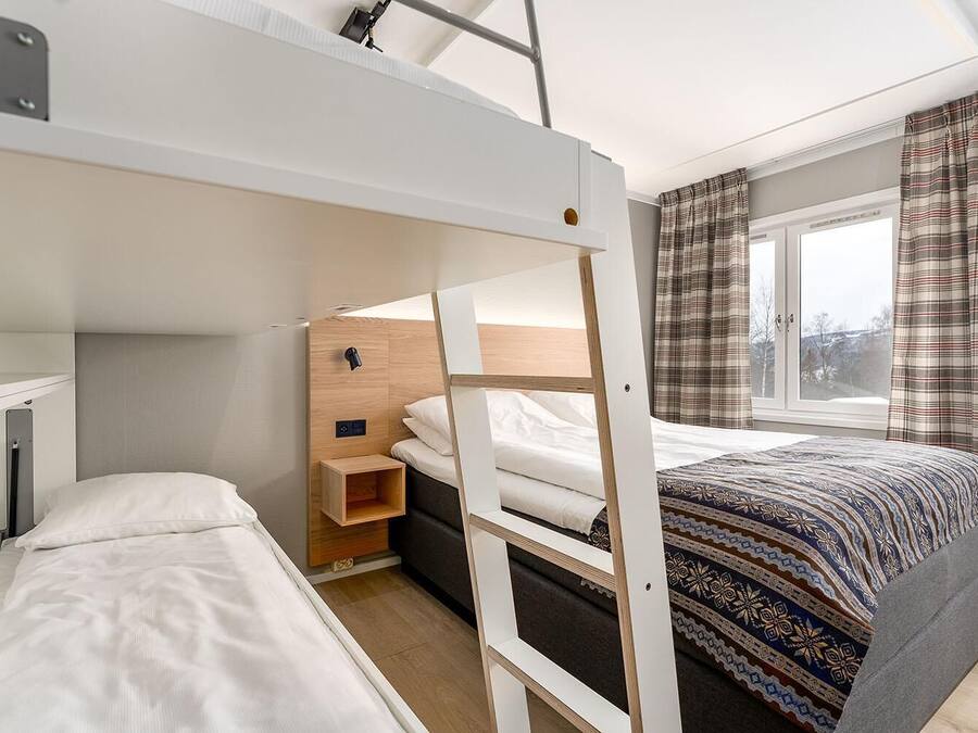 A compact hotel room with a bunk bed, a small bedside table, a window with curtains, and a patterned bedspread.