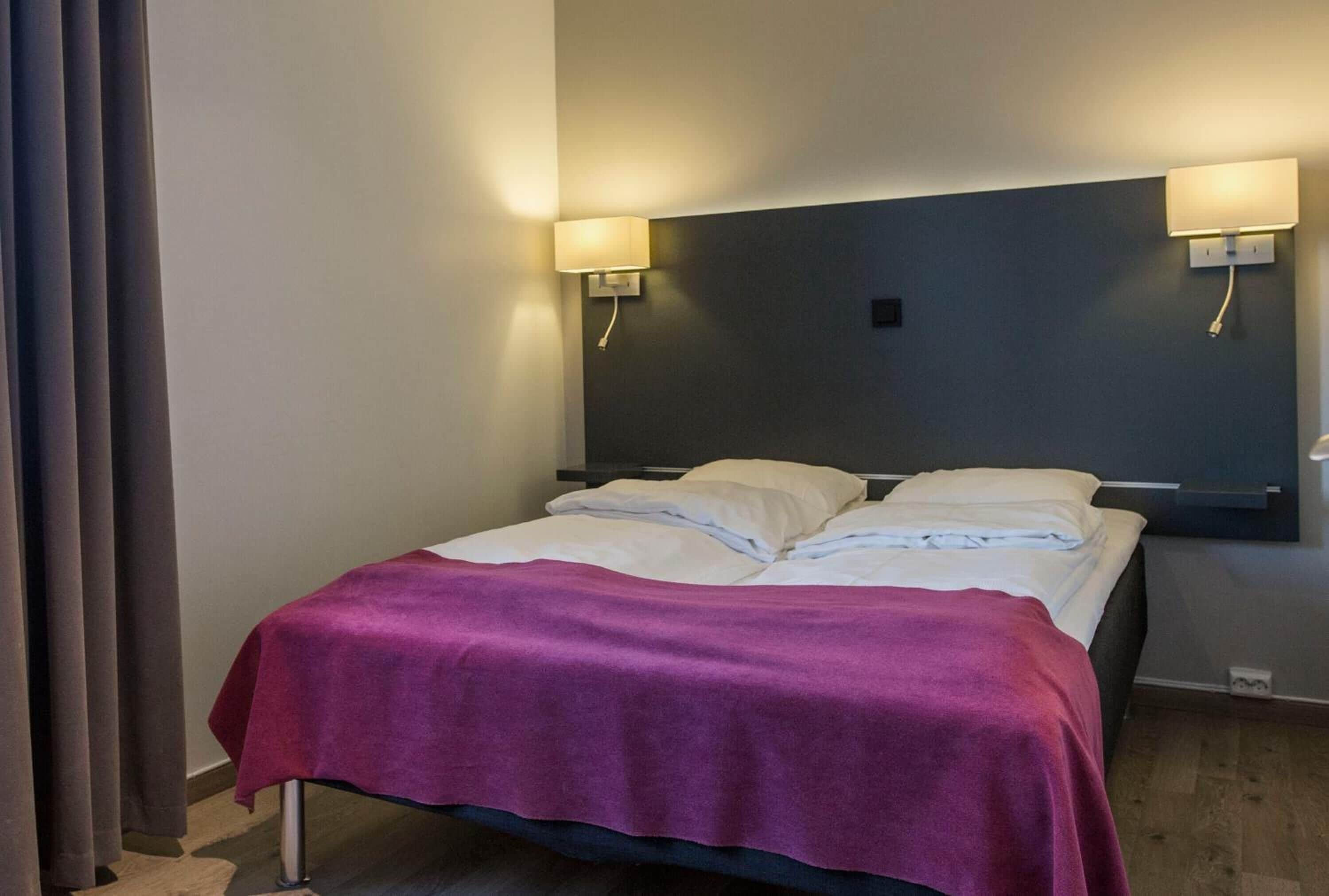 A single bed with a purple blanket, two wall-mounted lamps, and a dark headboard in a hotel room.