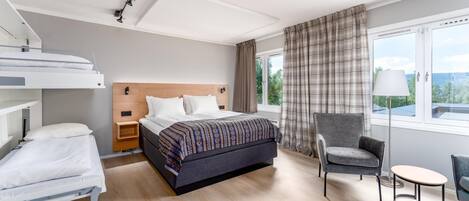 A hotel room with a bunk bed, a single bed, a chair, a small table, and a window with curtains.