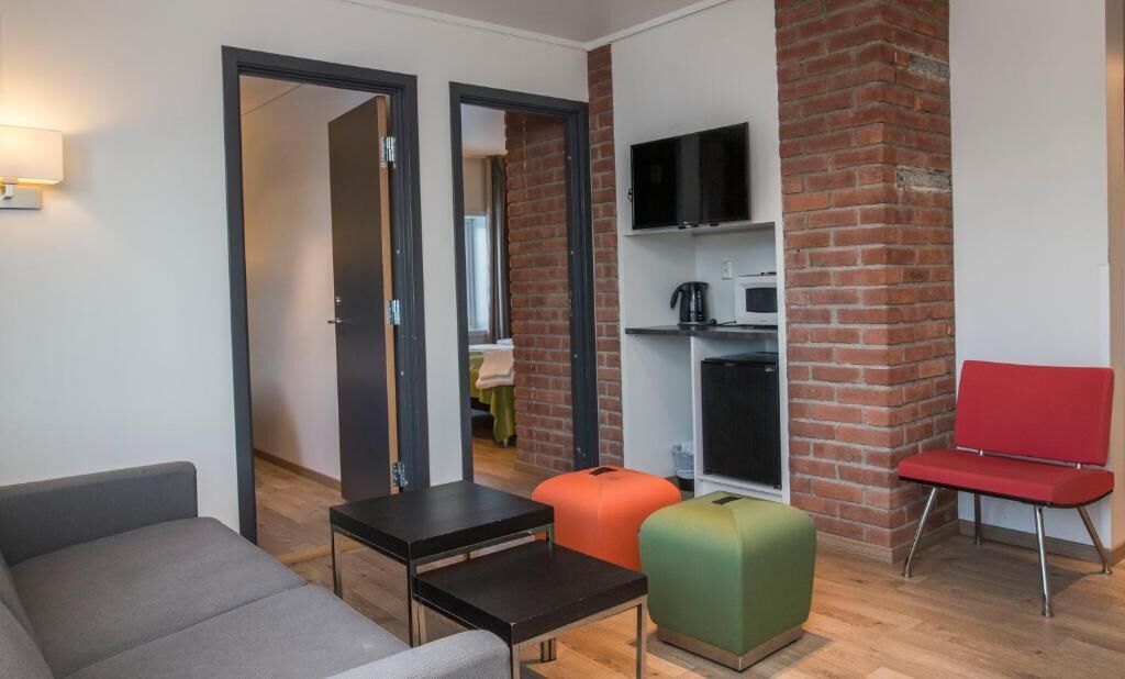 A modern living room with a sofa, a small table, a red chair, and a green ottoman. There is a television mounted on the wall and a brick column.