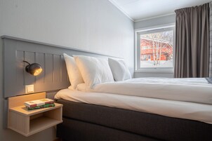Una cama bien hecha con sábanas blancas, una lámpara de noche y una mesita de madera con libros.