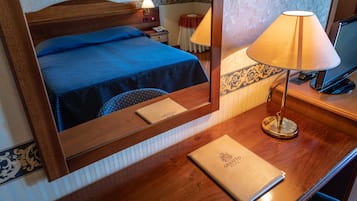 Een hotelkamer met een houten hoofdbord, een bed met een blauwe spreideken, een nachtkastje met een lamp, een spiegel en een klein tafeltje met een boek.