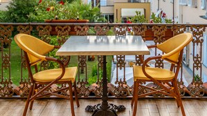 Un balcón con mesa y sillas, rodeado de barandales decorativos y vistas a un jardín y edificios.