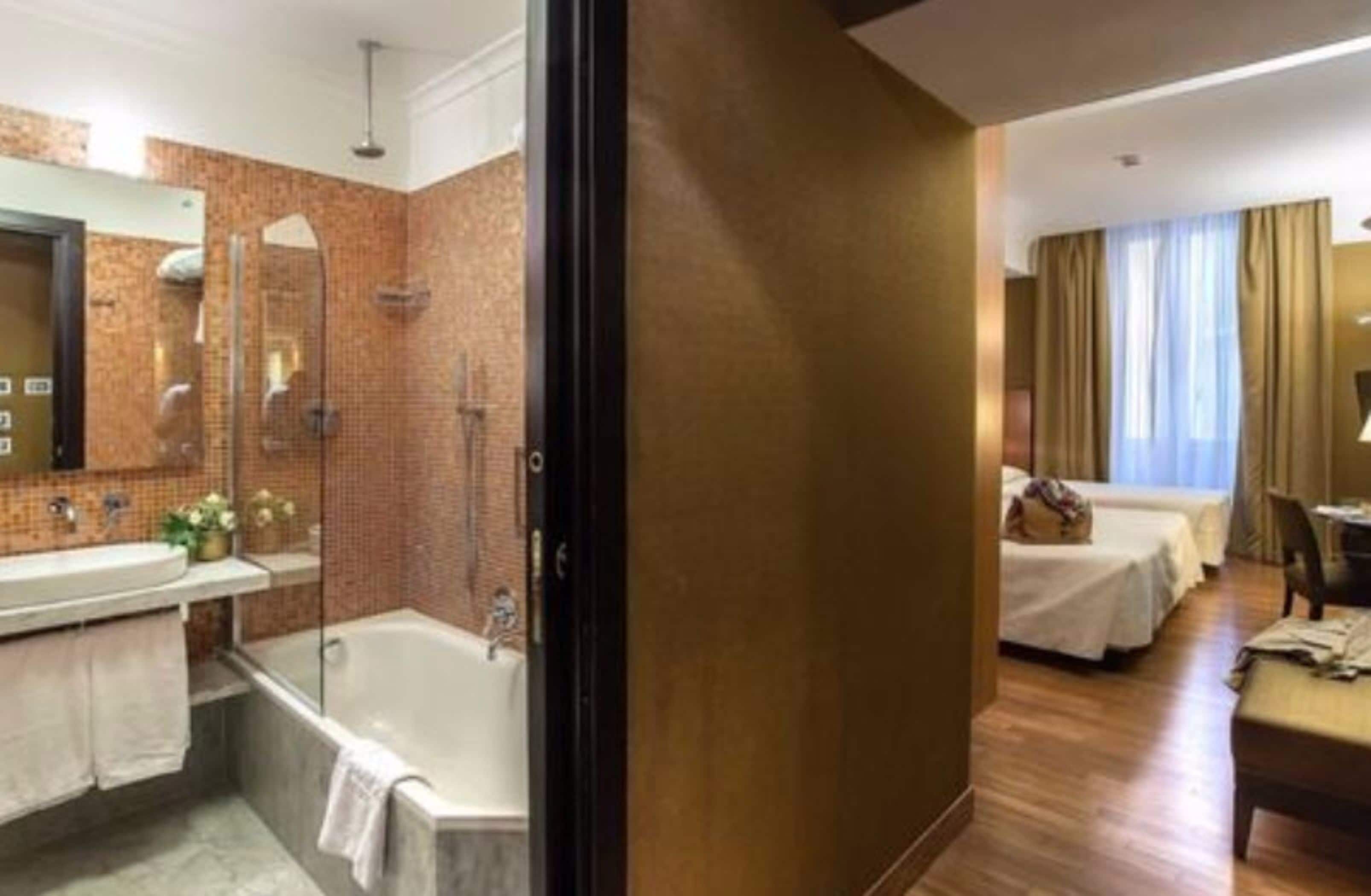 A hotel room with a bathroom featuring a bathtub, a shower, and a vanity with a mirror. There are two beds in the room.