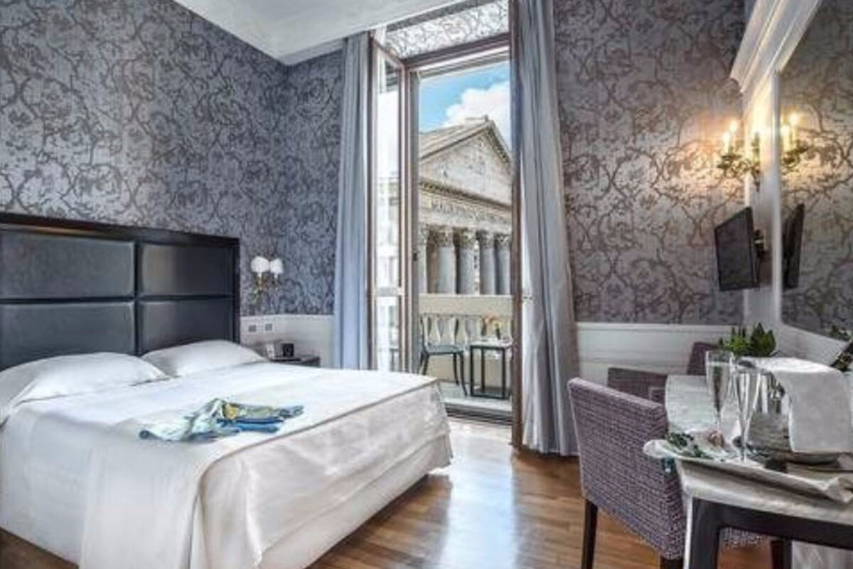 Una camera d'albergo con un letto grande, una zona pranzo con tavolo e sedie, e vista su architettura classica attraverso una finestra.