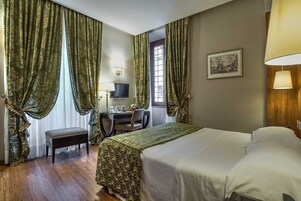 A hotel room with a bed, a desk with a computer, a chair, and a window with curtains.