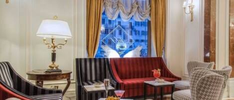 A hotel room with a red sofa, striped armchair, and patterned armchair. There is a small table with a drink and snacks, and a lamp on a side table.