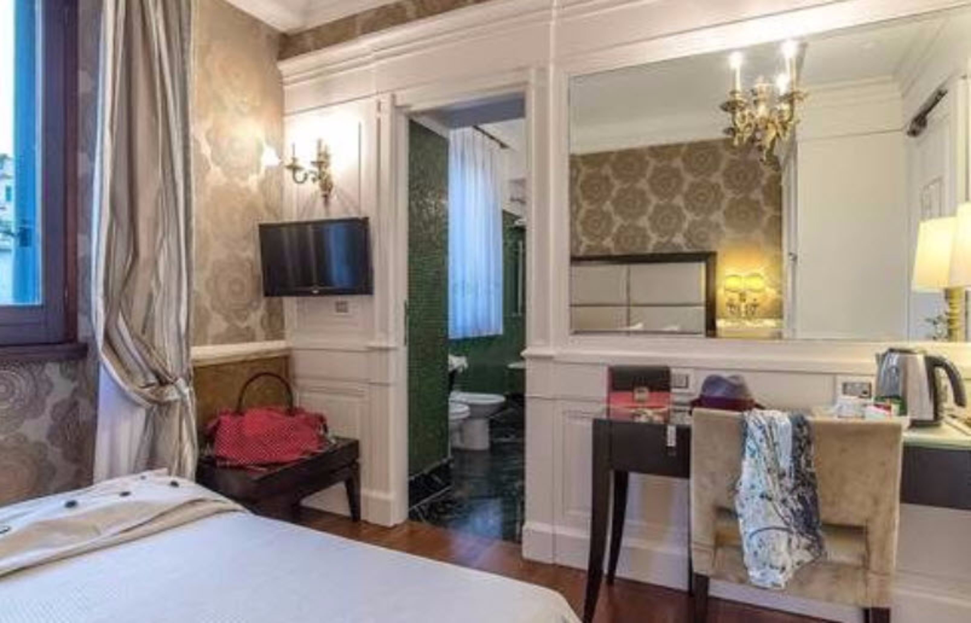 A hotel room with a bed, a desk, a chair, a TV, a mirror, a window with curtains, and a bathroom visible through an open door.
