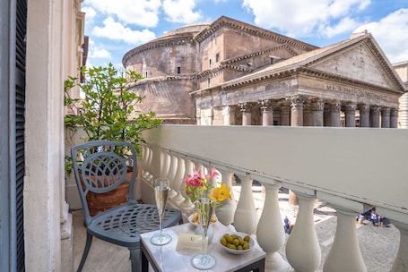 A balcony with a table, two chairs, a vase with flowers, a bowl of fruit, and a view of ancient Roman architecture.