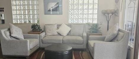 A living room with a grey sectional sofa, a round coffee table, and two side tables with plants. There are two windows with patterned curtains, a painting on the wall, and a floor lamp.