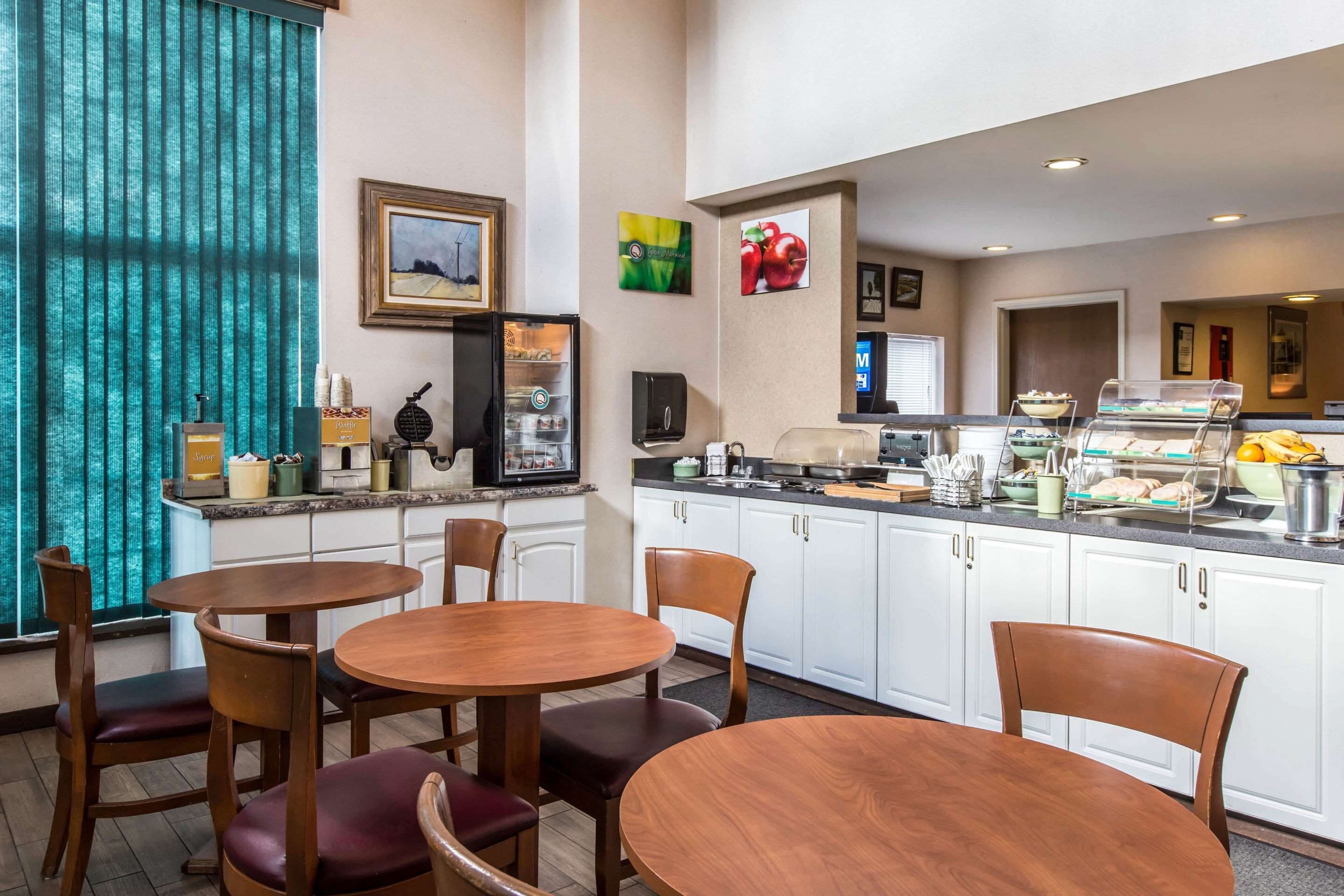 Une cuisine d’hôtel avec un buffet, comprenant une machine à café, un réfrigérateur et divers aliments. Des tables et des chaises en bois sont disposées pour les repas.