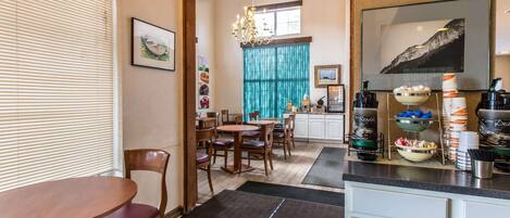 A dining area with a wooden table and chairs, a chandelier, and a kitchen area with a coffee station.