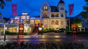 A historic building with a grand entrance, multiple flags, and a well-lit facade. - Hotel IV Jahreszeiten Kühlungsborn  (Kuehlungsborn)
