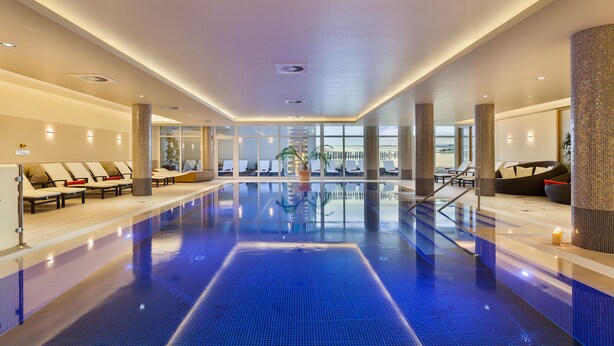 A large indoor swimming pool with clear blue water, surrounded by lounge chairs and a modern architectural design. - Hotel IV Jahreszeiten Kühlungsborn  (Kuehlungsborn)
