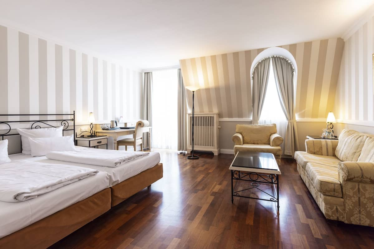 Quarto de hotel com duas camas, uma escrivaninha, uma cadeira, um sofá e um abajur.