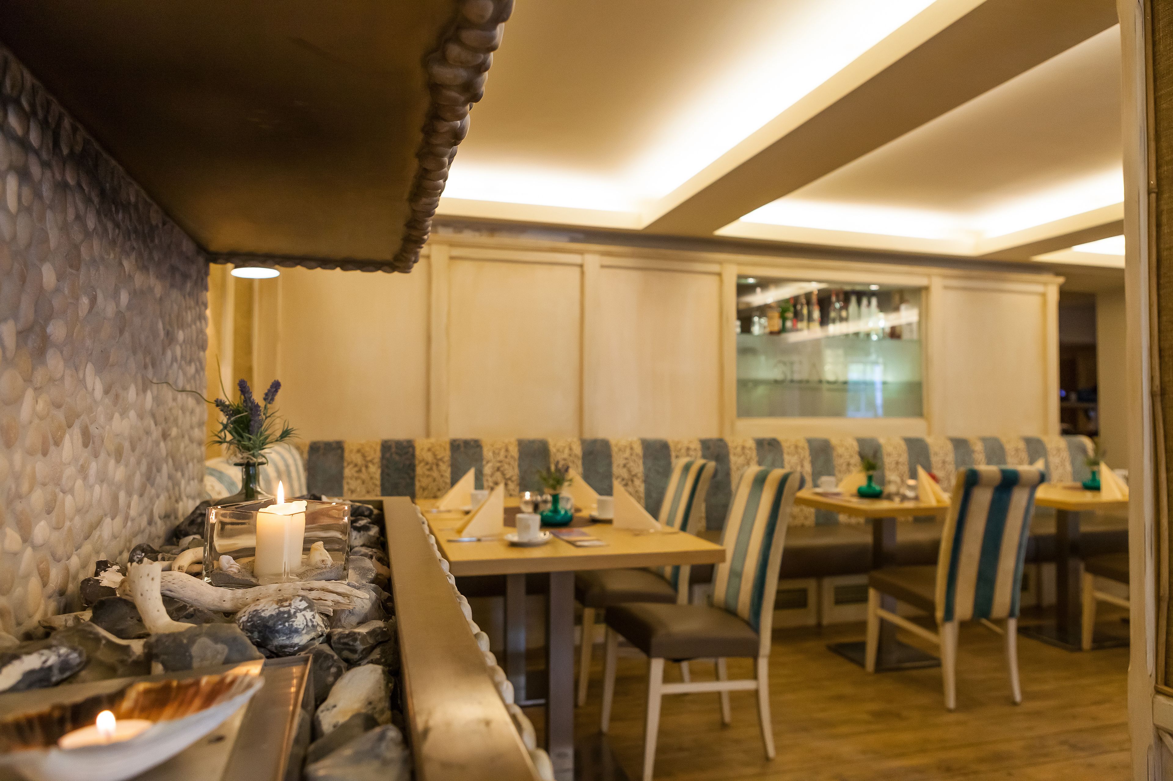 Ein Restaurant mit Sitzboxen und Tischen, eine Bar im Hintergrund sowie eine dekorative Tischdekoration mit Kerzen.