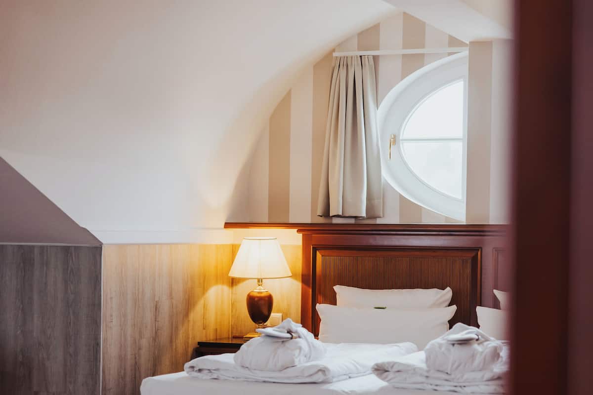 Quarto de hotel com piso de madeira, uma cama com lençóis brancos, um banco, mesa de cabeceira com abajur e janela com cortinas.