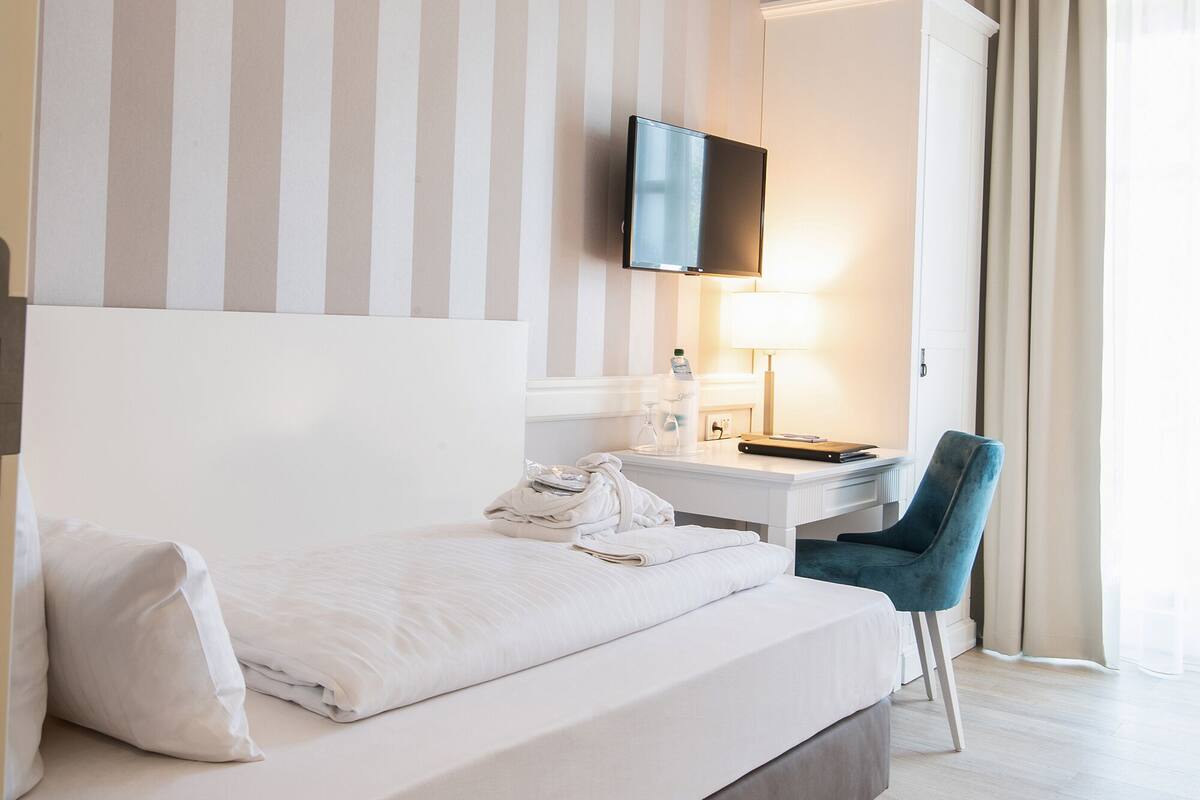 Quarto de hotel com uma cama grande, uma escrivaninha com cadeira, televisão na parede e uma janela com cortinas.
