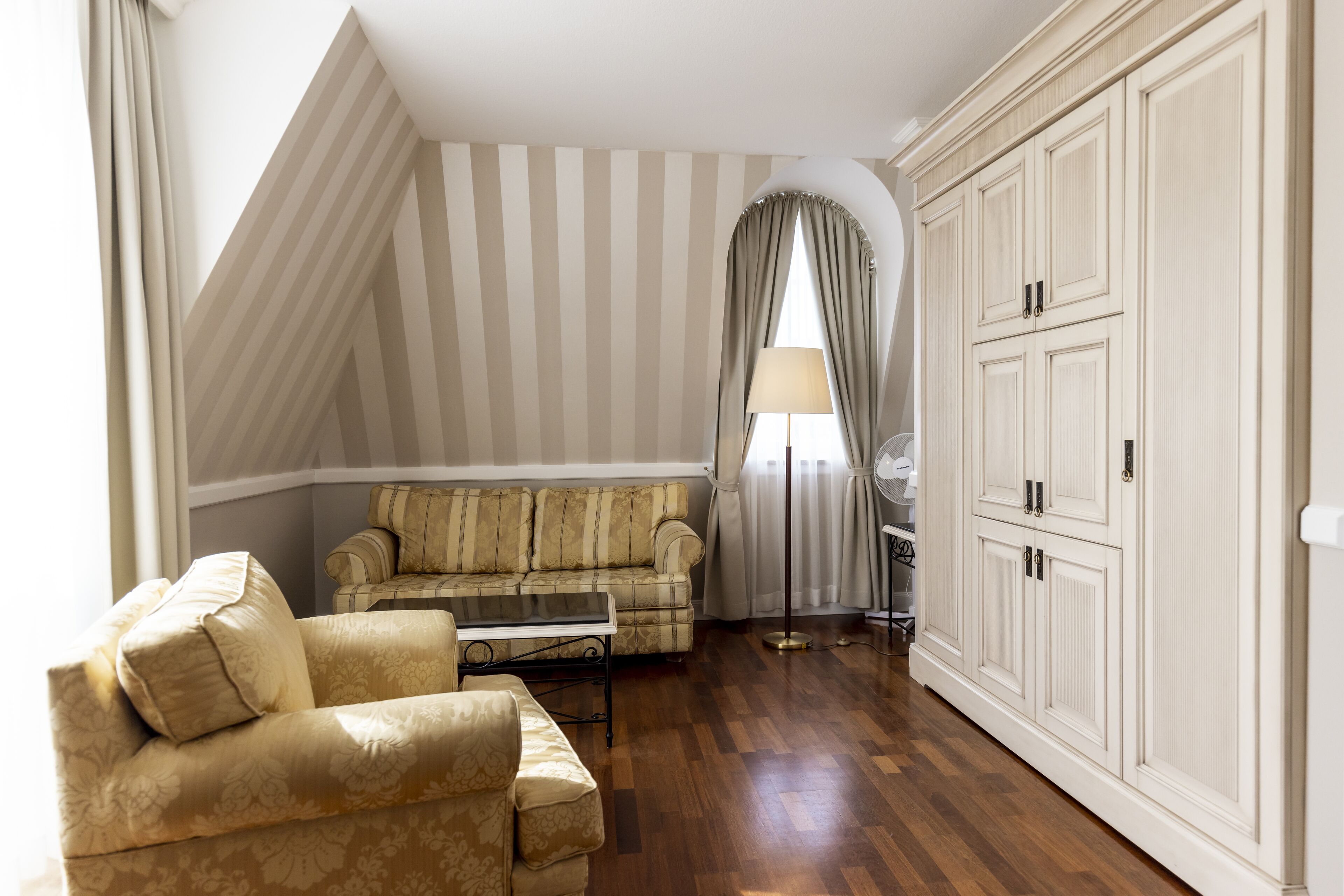 A room with a sofa, armchair, and a large arched window with curtains.