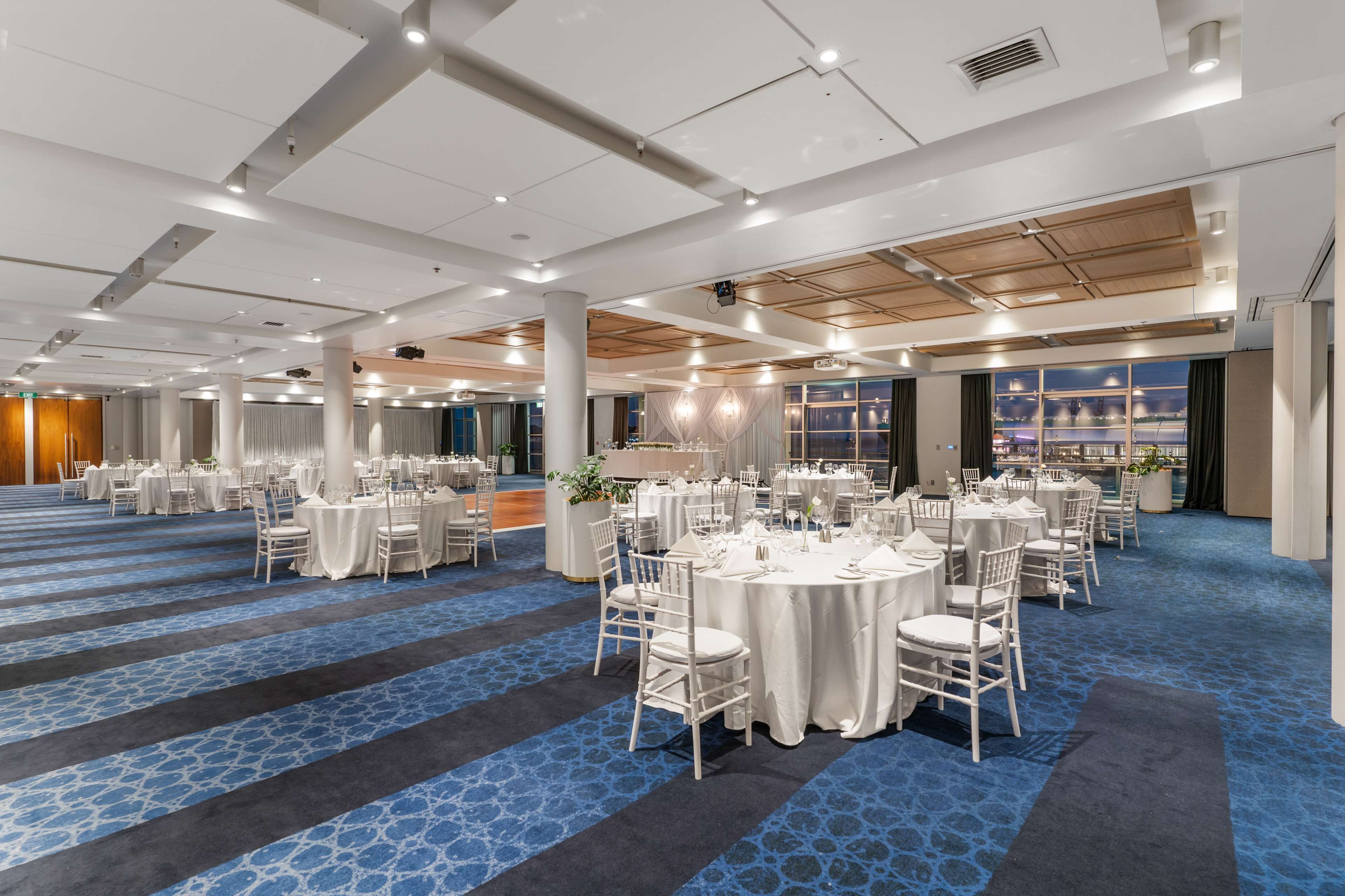A large banquet hall with round tables and chairs, a patterned carpet, and a high ceiling with recessed lighting.
