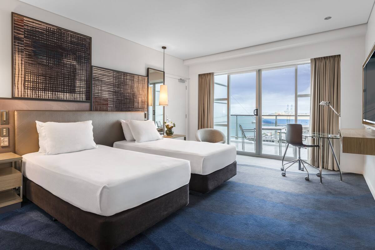 A hotel room with two beds, a desk, a chair, a TV, and a balcony with a view of the sea and a bridge.
