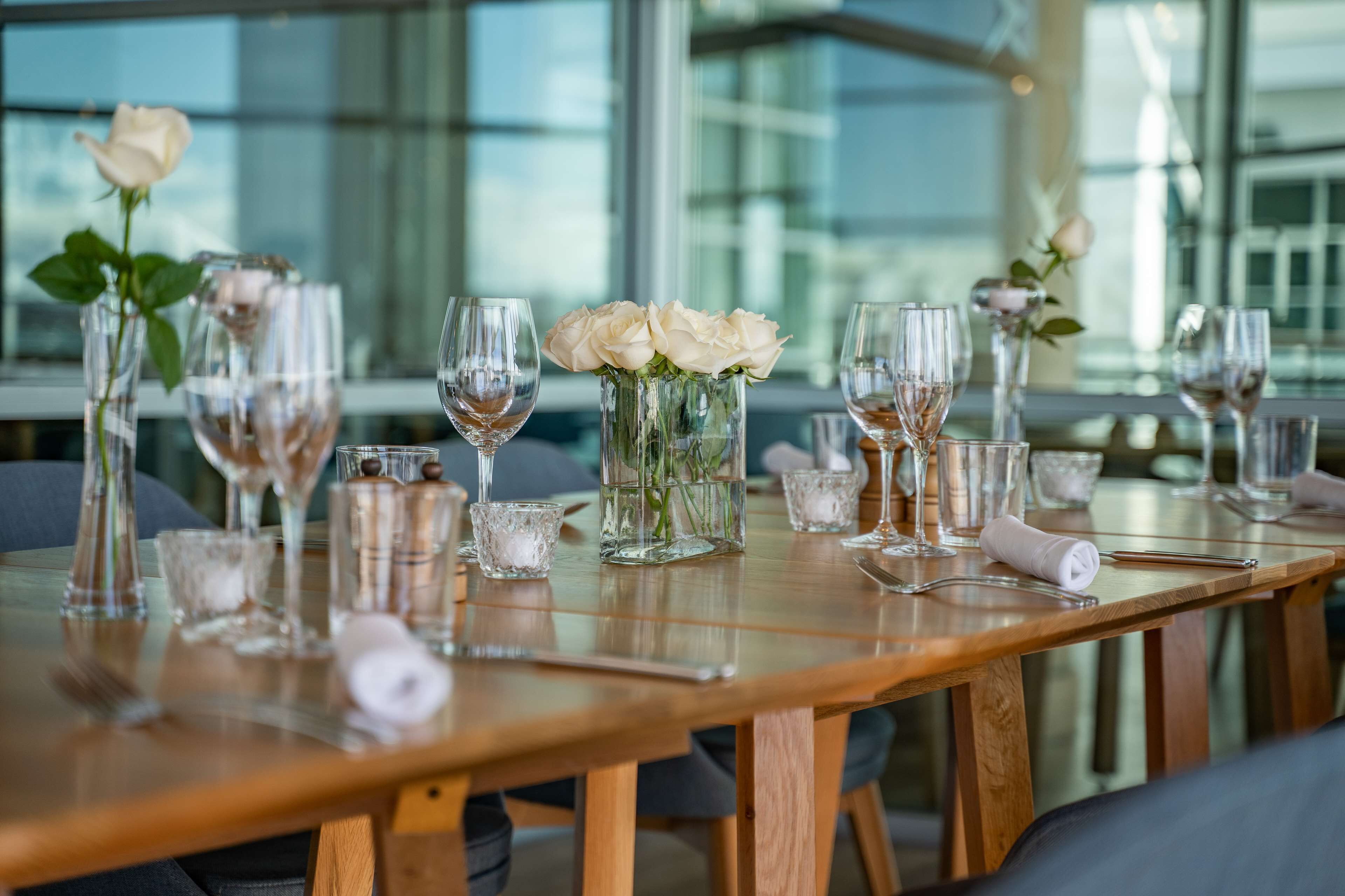 A dining table set with wine glasses, a vase with white roses, and a rolled napkin.