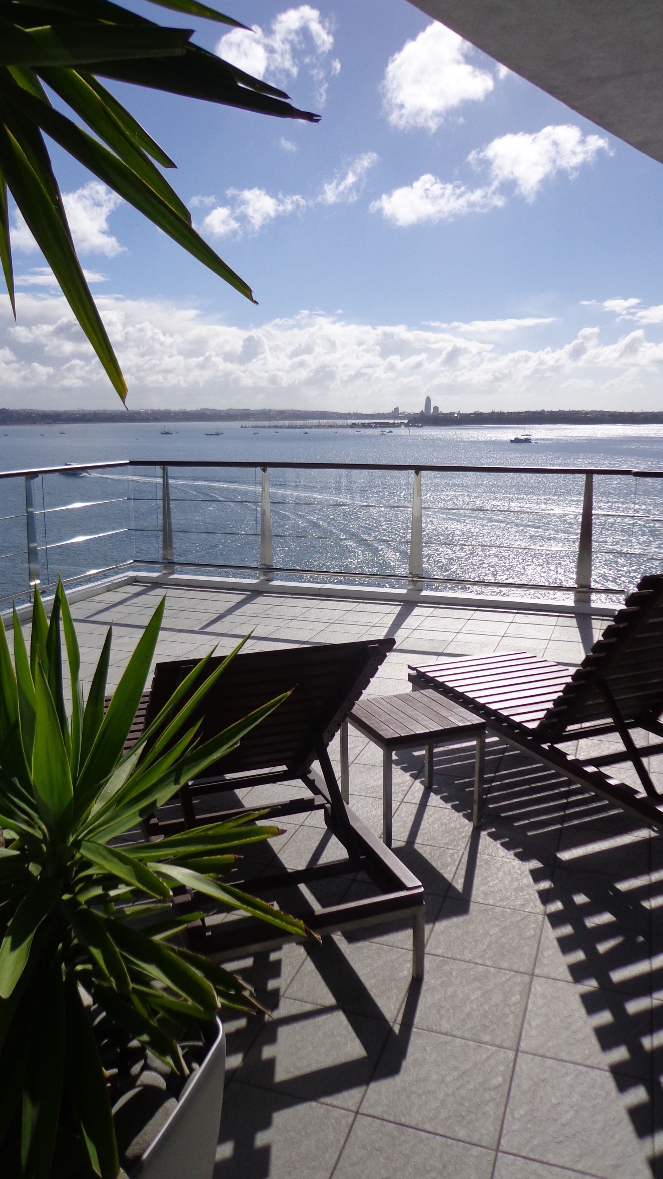 A terrace with a view of the sea, featuring a railing, two lounge chairs, and a potted plant.