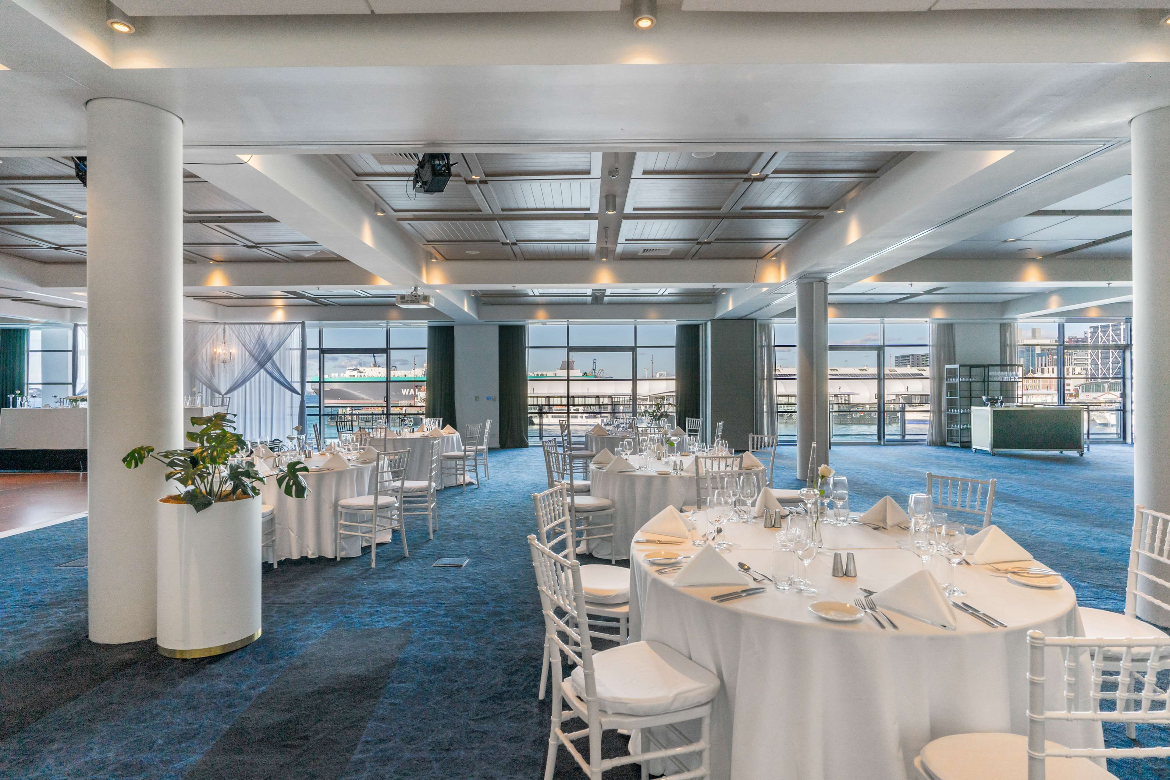 Un amplio salón de banquetes con mesas redondas dispuestas para cenar, grandes ventanales con vistas a la ciudad y un techo alto con vigas a la vista.