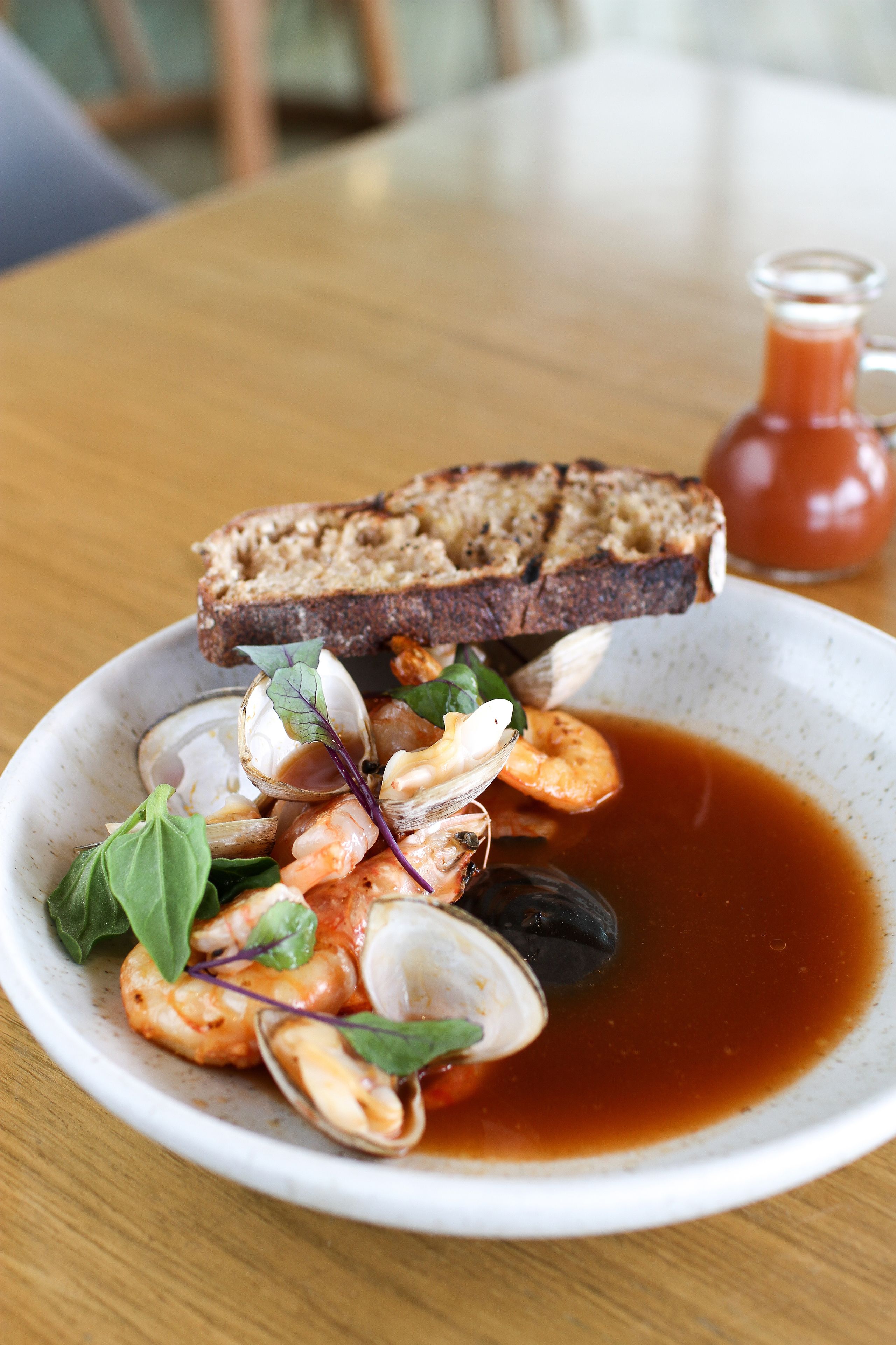 A bowl of seafood soup with clams and shrimp, accompanied by a slice of toasted bread and a small glass of tomato sauce.