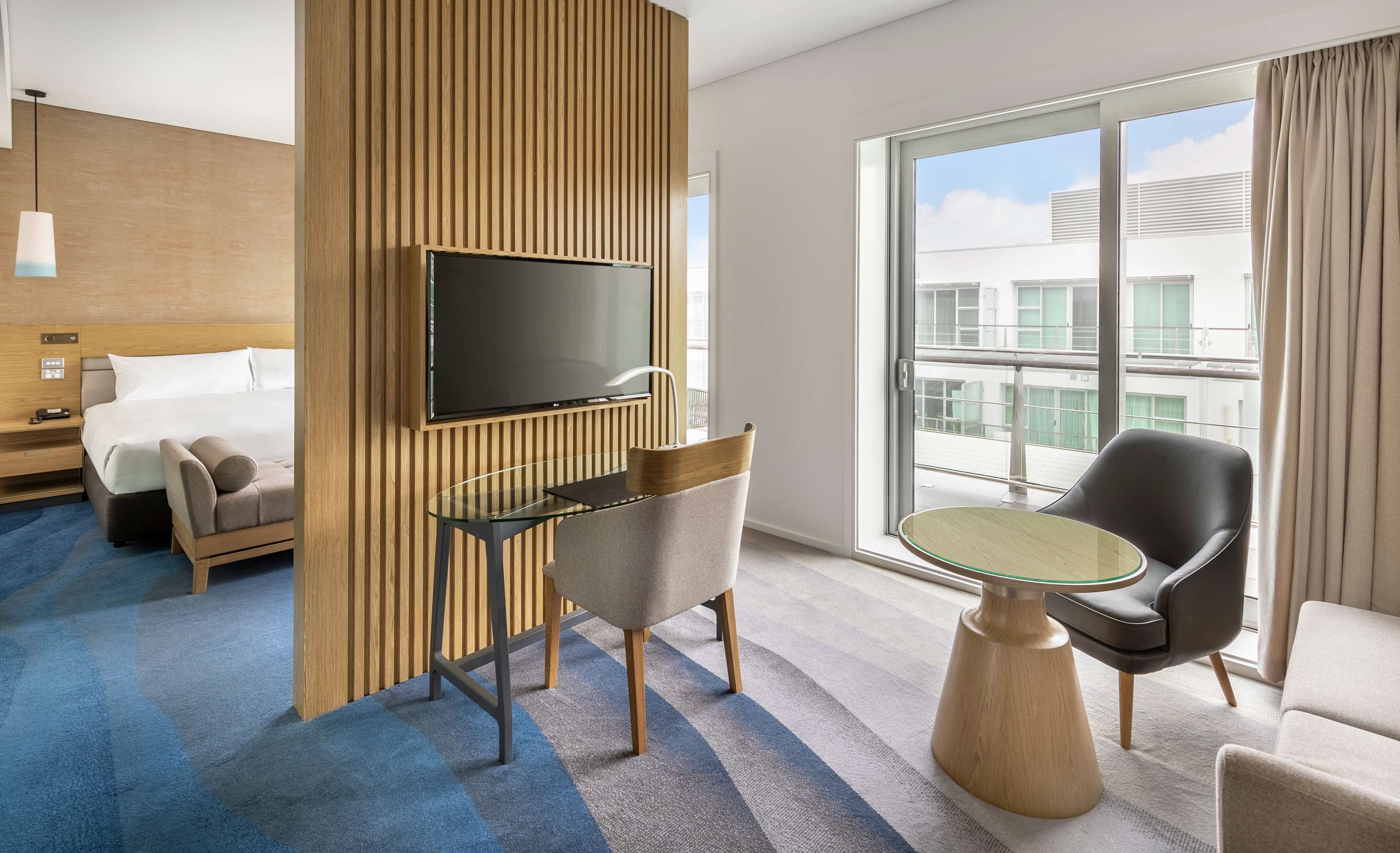 A hotel room with a bed, a TV mounted on a wooden panel, a glass table, two chairs, and a large window with a city view.