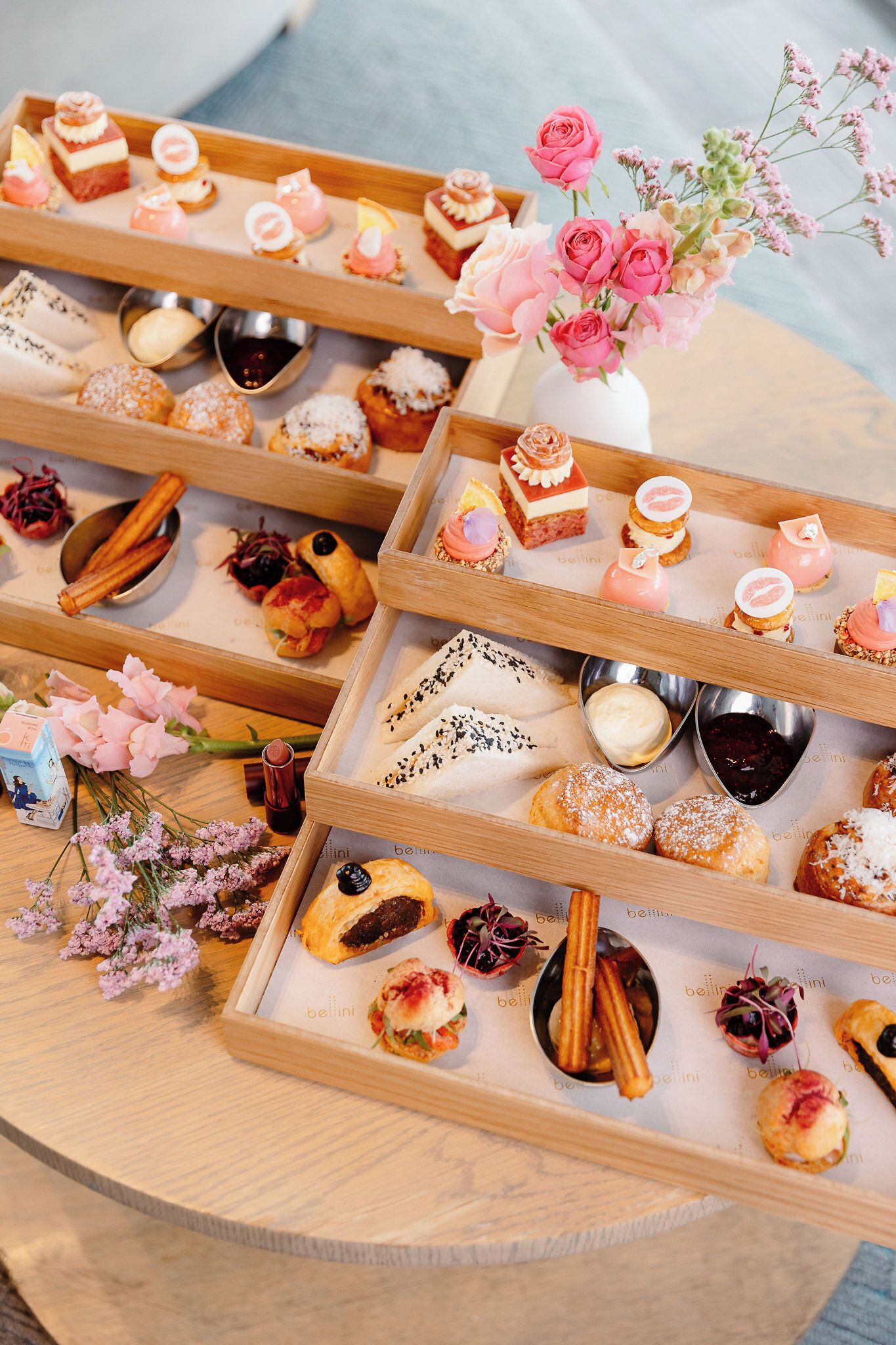 Diverse gebakjes en desserts, gerangschikt op houten schalen, met kleine kommetjes met saus en een vaas roze bloemen.