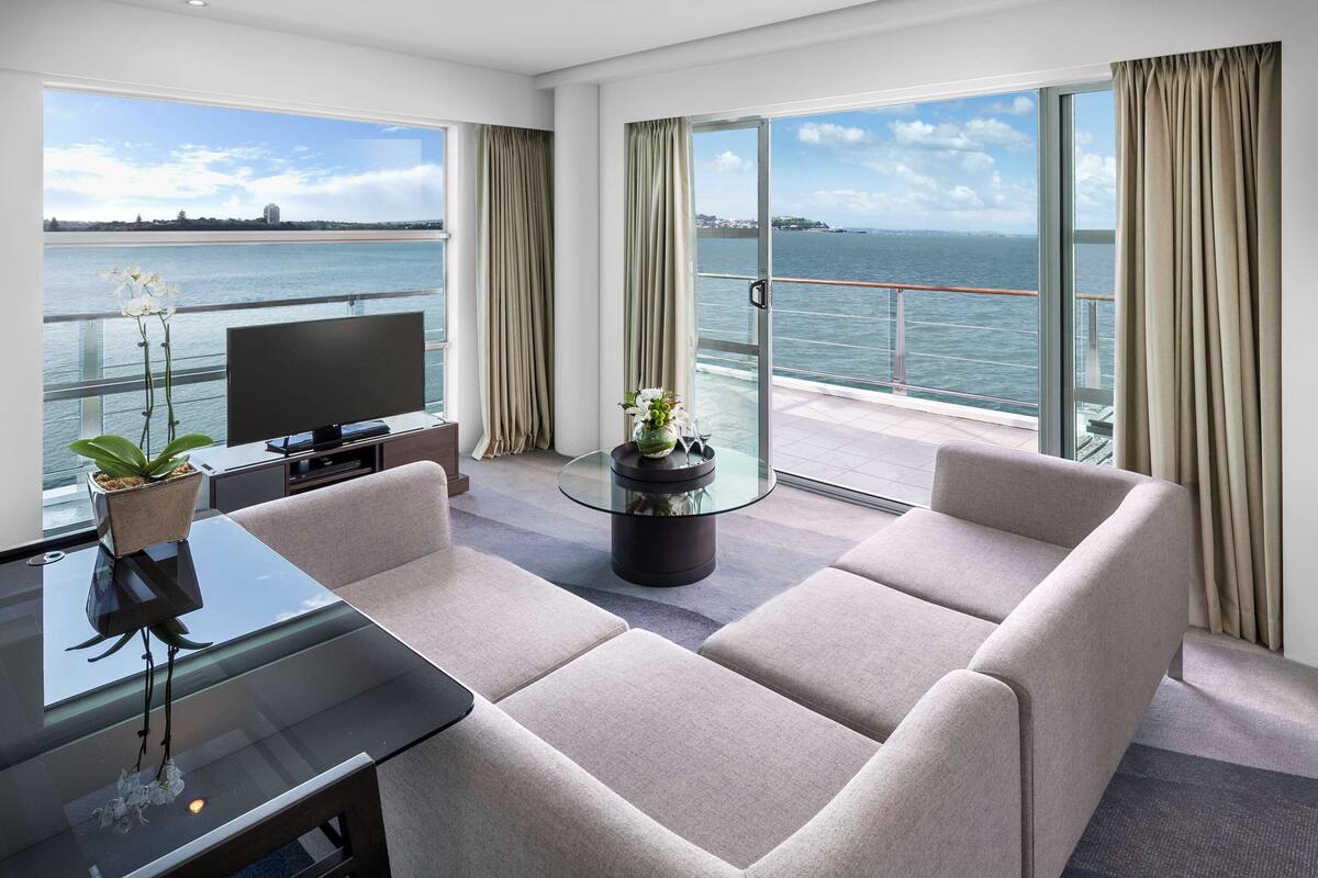 A modern hotel room with a large balcony, a glass table, a sofa, and a TV.