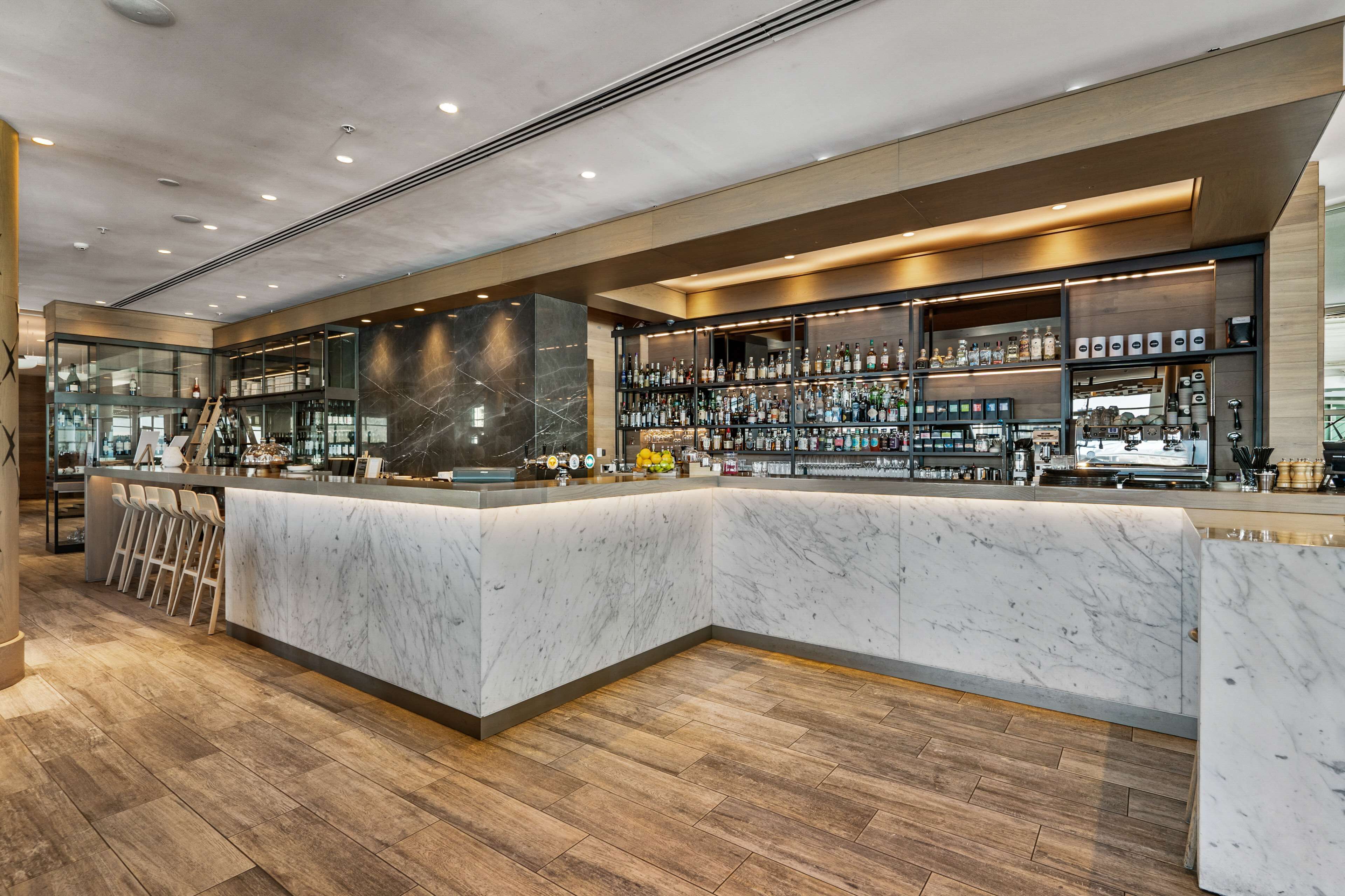 A modern bar area with a marble counter, wooden flooring, and shelves stocked with bottles.
