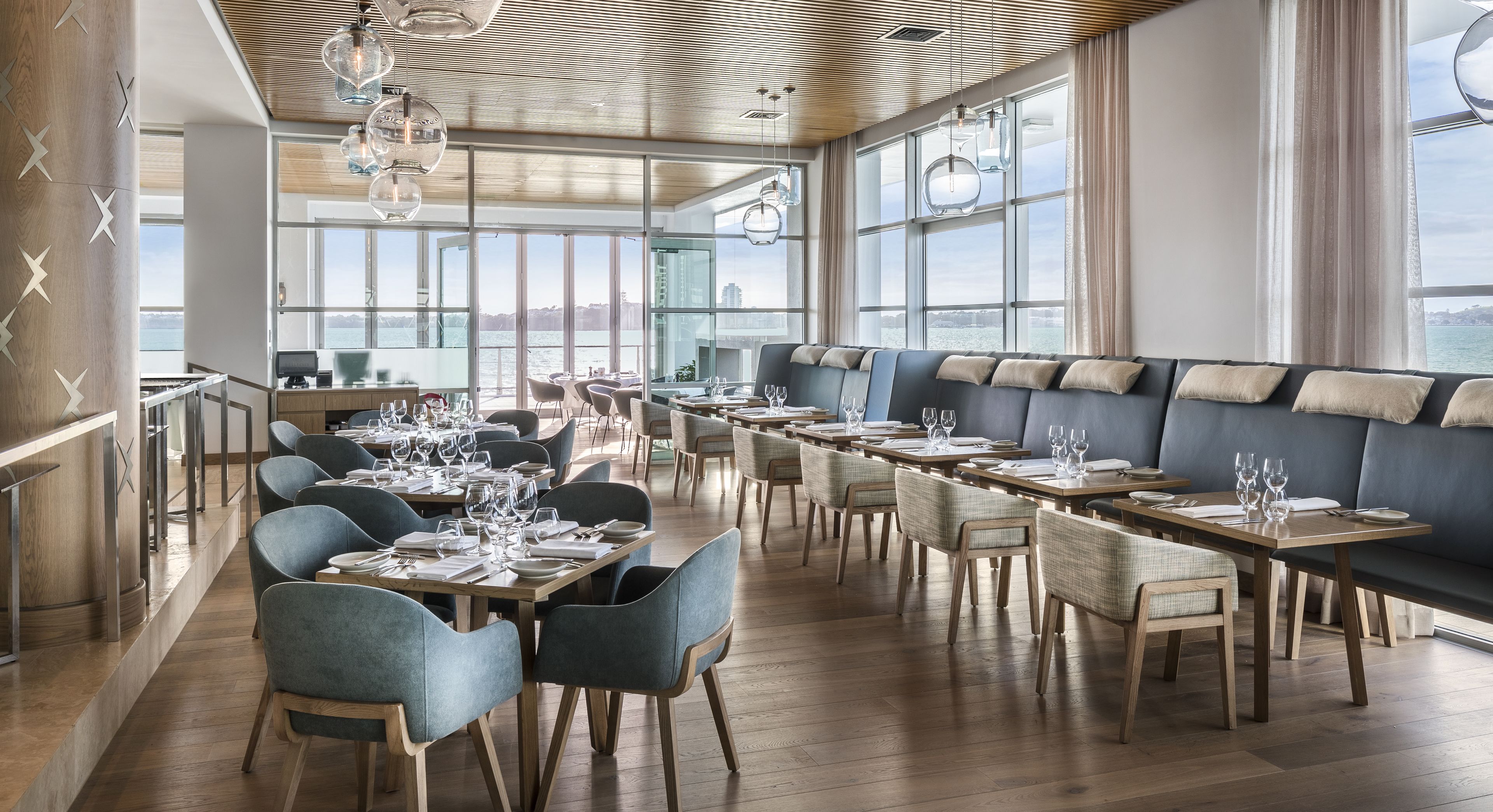 A modern restaurant with a long dining area, wooden flooring, and large windows offering a view of the city.
