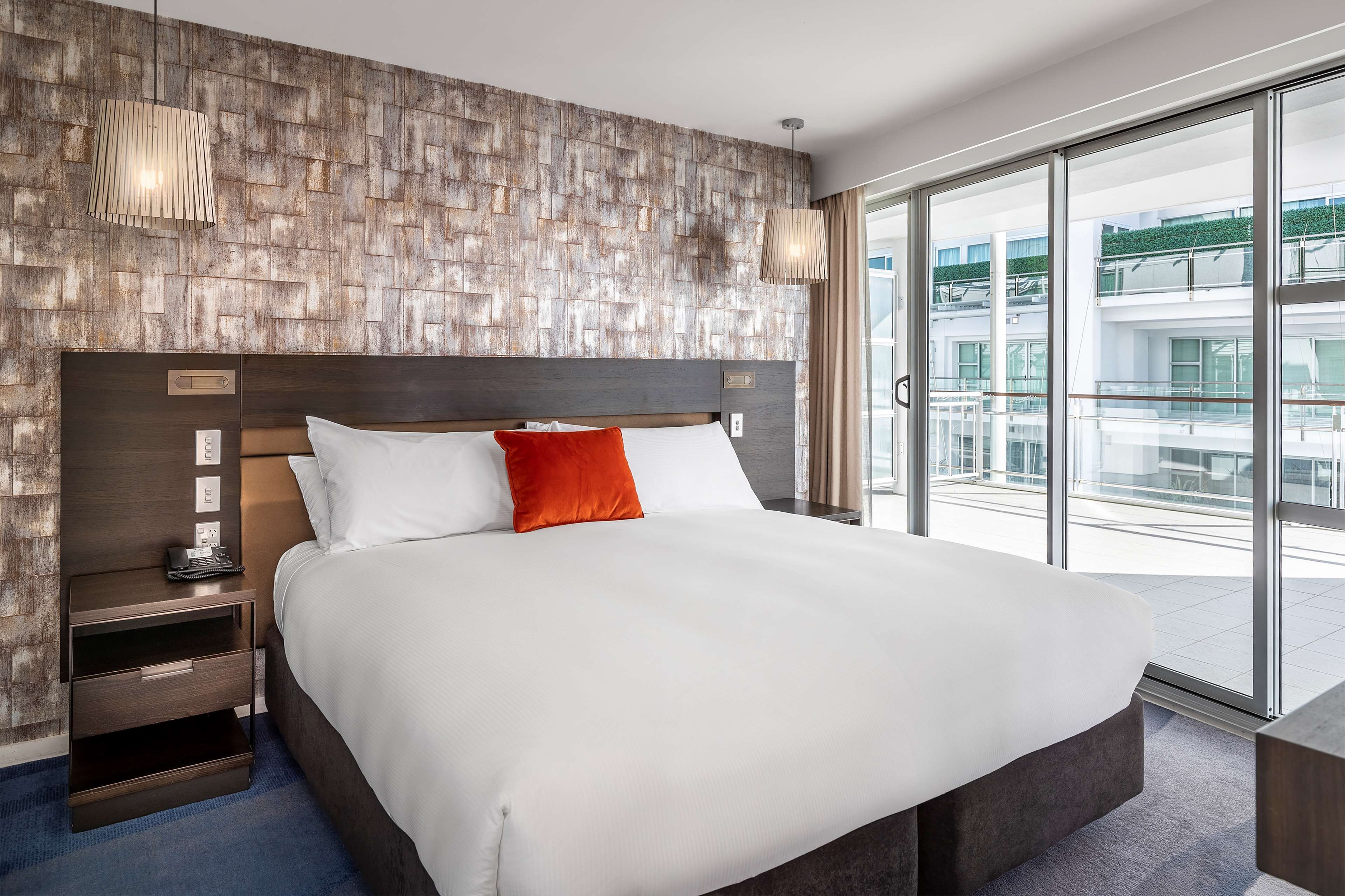 A modern hotel room with a large bed, a wooden headboard, a nightstand, and a view of the city through large windows.
