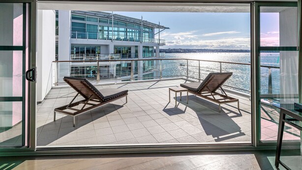 Un balcon doté de deux chaises longues et d’une balustrade en verre, offrant une vue sur une étendue d’eau et un bâtiment moderne.
