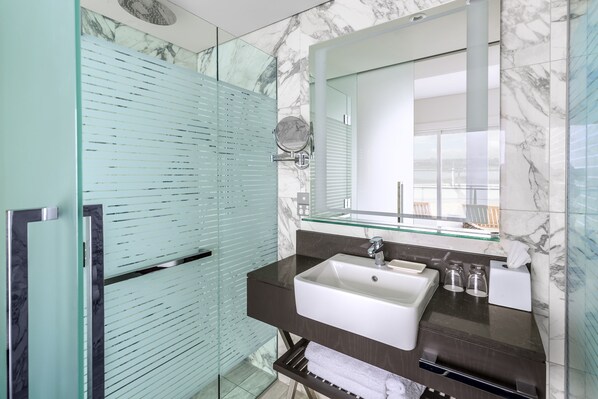 A modern bathroom with a glass shower enclosure, a marble-tiled wall, a large mirror, and a dark countertop with a sink.