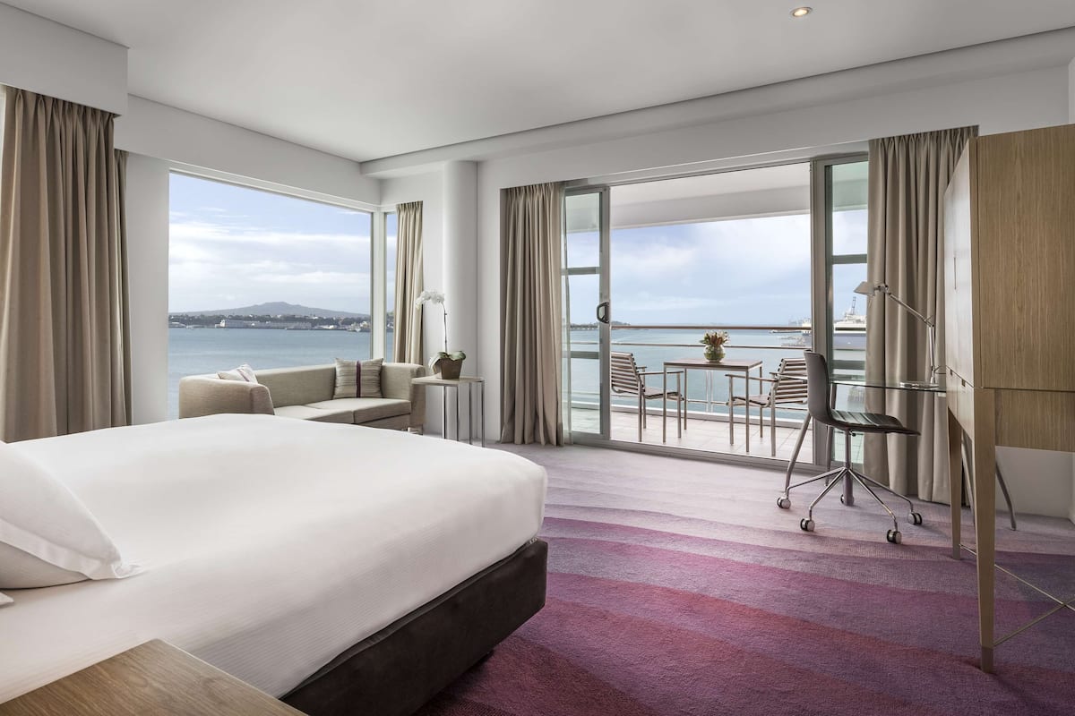 A modern hotel room with a large bed, a balcony with a table and chairs, and a view of the sea and city skyline.