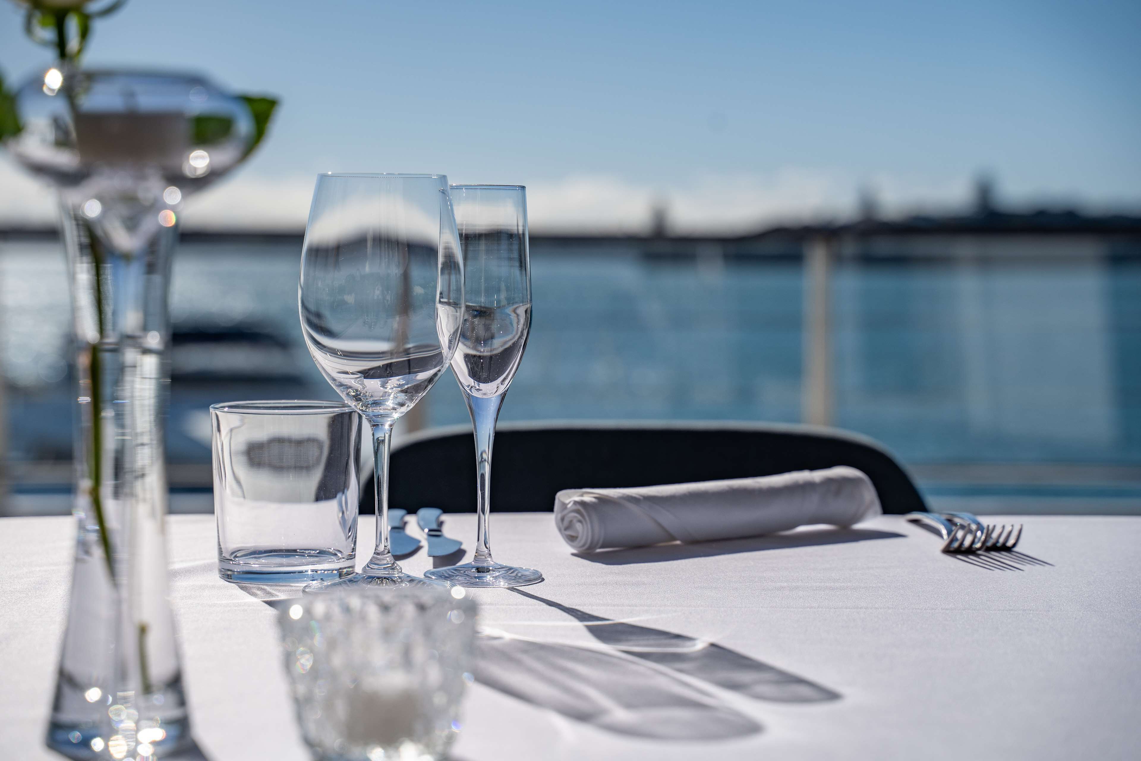 A table set with wine glasses, a water glass, a fork, and a knife, with a view of the water in the background.