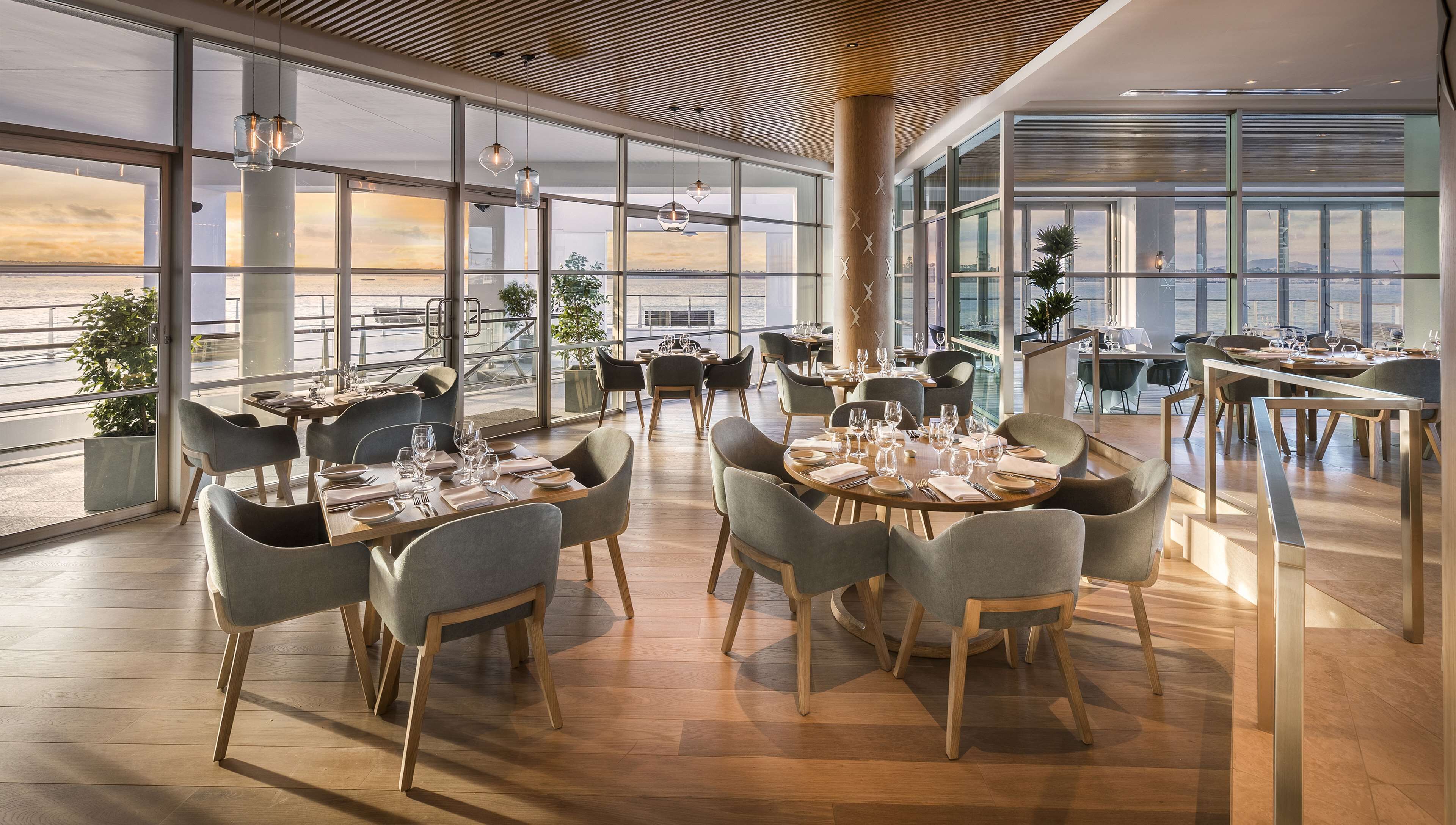 A modern restaurant with large windows offering city views, wooden flooring, and contemporary furniture.